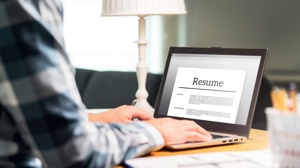 a man sits at his desk to type a two-page resume on his laptop